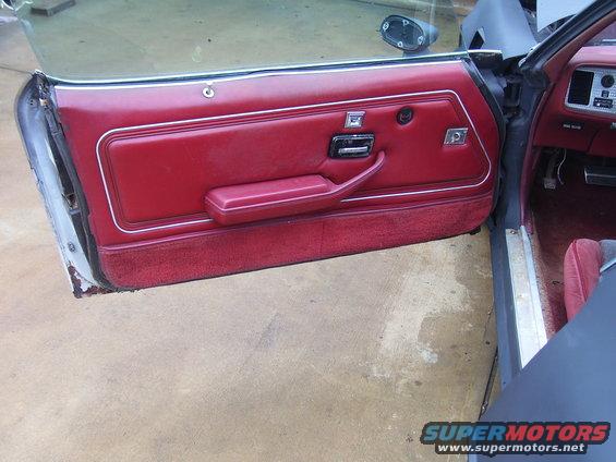 Interior driver's side door panel with red upholstery and chrome accents. Car is likely a 1970s or 1980s model