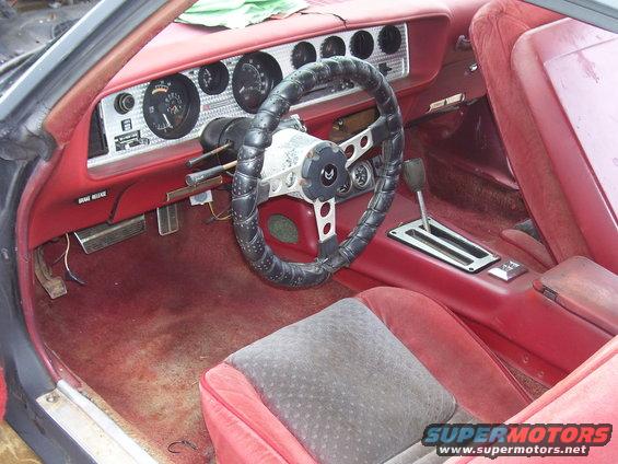 Car interior with red seats, dashboard, and steering wheel. The floor is worn and dirty