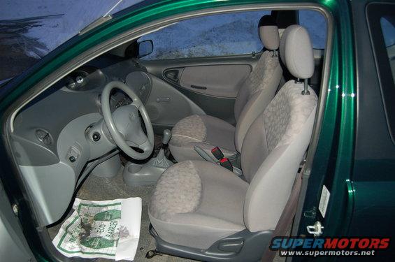 Green car interior showing front seats, dashboard, and steering wheel. Paper on floor has 'ECO BARRIER' text