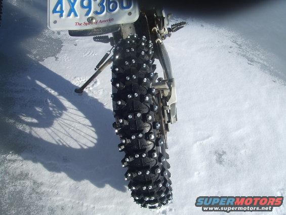 Rear tire of a motorcycle with studs for snow, parked on a snowy surface. License plate visible