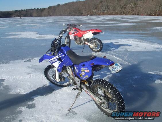 Blue Yamaha dirt bike with studded tires on ice, red Honda in background