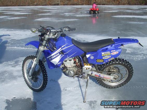 Blue Yamaha dirt bike with studded tires on ice, ATV in background