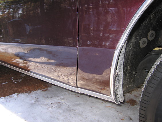 Close-up of a dark red car's lower door panel and front wheel well with chrome trim