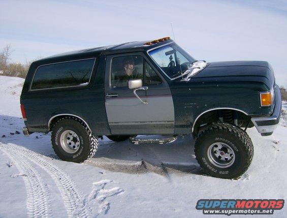 Dark green and gray Ford Bronco with aftermarket wheels and tires in the snow. Driver is visible inside