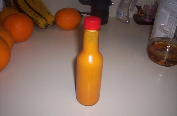 Bottle of orange sauce with a red cap sits on a counter next to oranges and bananas