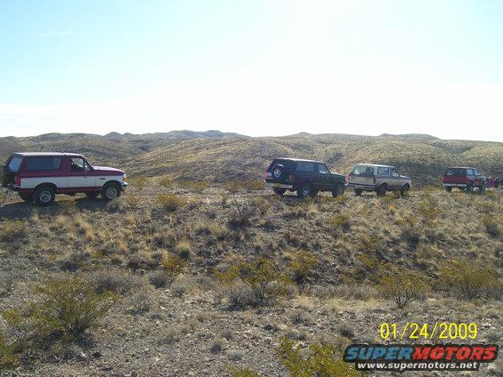 Four Ford Broncos parked on a dry, hilly landscape under a clear sky. The lead Bronco is two-tone red and white