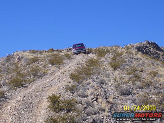 Red SUV driving up a rocky, arid hill under a clear blue sky