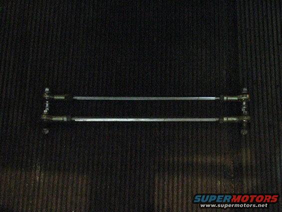 Two metal tie rods with threaded ends and ball joints, laid horizontally against a dark, textured surface