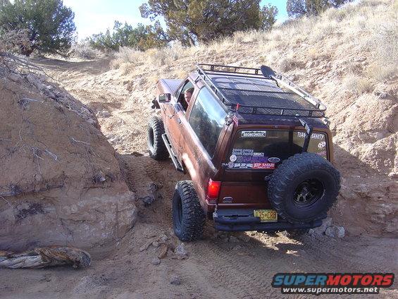 Brown Ford Bronco off-roading on a dirt hill, with a spare tire mounted on the rear and a roof rack