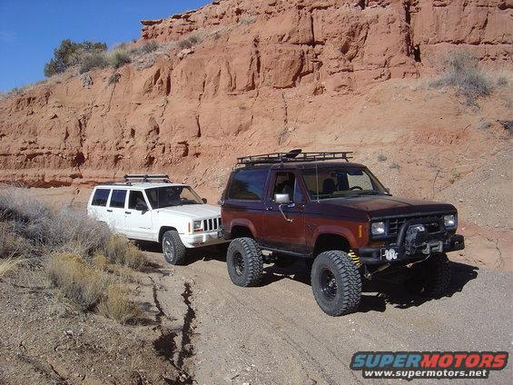 Brown SUV and white Jeep Cherokee on a dirt road with red rock cliffs