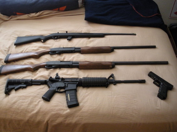 Four firearms, including an AR-15 style rifle, two shotguns, and a handgun, are arranged on a bed