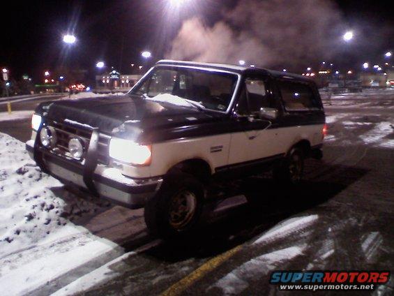 Two-tone Ford Bronco parked in a snowy lot at night, exhaust visible