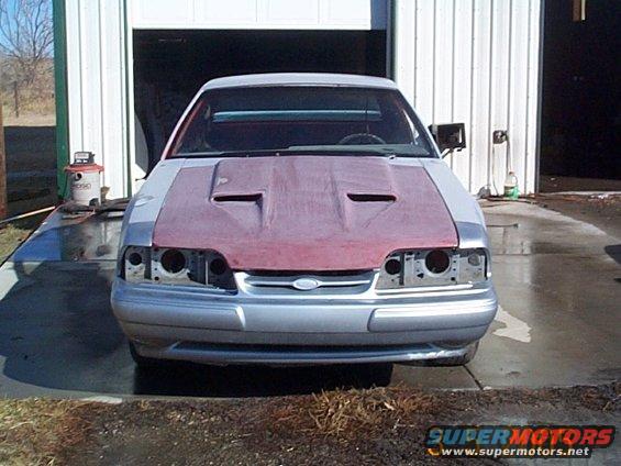 Front view of a silver Ford Mustang with a red hood and no headlights, parked in front of a garage