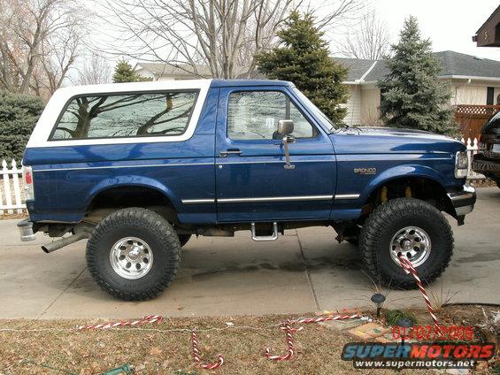 Blue Ford Bronco with a white hardtop, lifted suspension, and large tires, seen from the side