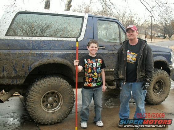 Muddy blue Ford Bronco with a boy holding a rake and a man standing next to it