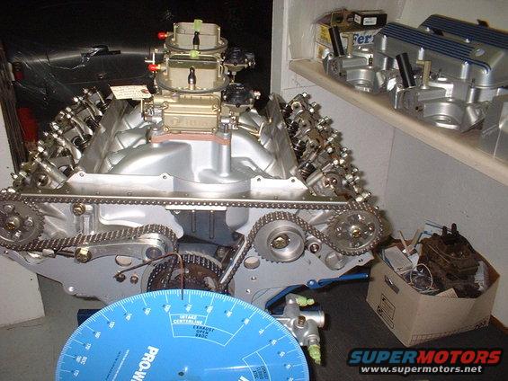 V8 engine block with dual carburetors, timing chain, and a blue degree wheel in foreground. Parts on shelf in background