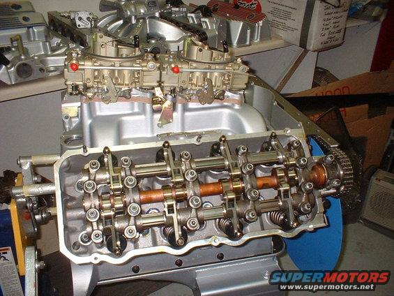 Close-up of a V8 engine block with valve train exposed, dual carburetors on top, and a timing chain visible