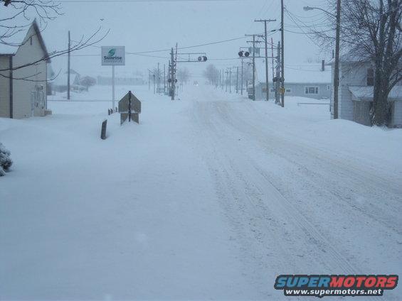 Snow-covered street with houses, power lines, and a sign visible