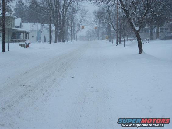snowy street with houses and trees, traffic lights visible