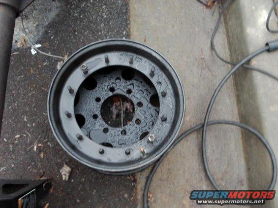 black spare tire rim with water droplets on it sits on the ground next to some cables