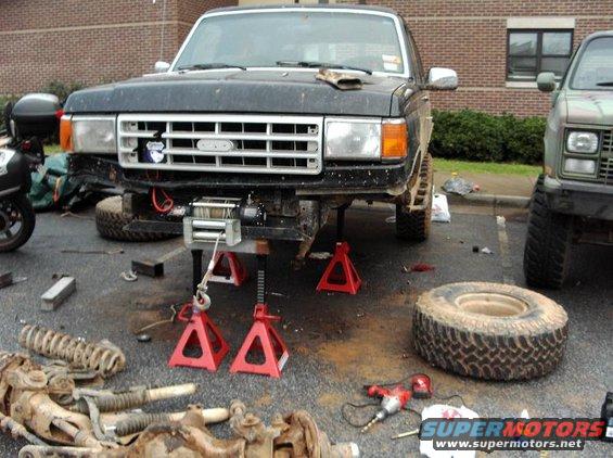 1990 Ford Bronco on jack stands with front end disassembled, another truck parked to the right