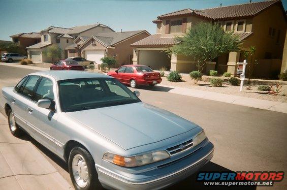 Silver sedan parked on street, front quarter view. Visible grille, headlights, and hood emblem. Houses in background