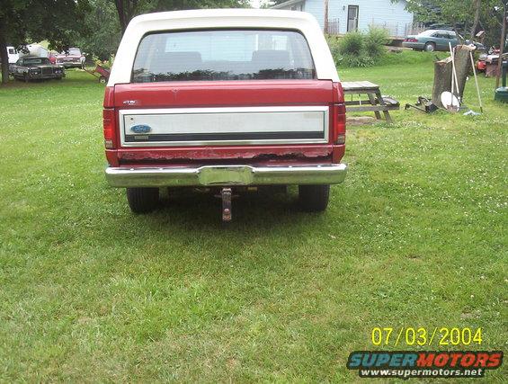 1985 Ford Bronco rear view, showing tailgate, bumper, and hitch receiver. Other vehicles and a picnic table are visible in the background