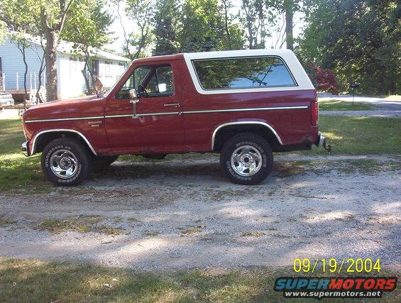 Red 1980s Ford Bronco SUV with a white removable top, parked on a gravel driveway