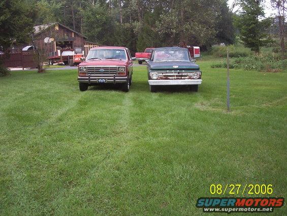 Red 1980s Ford Bronco and green 1960s Ford F-100 truck parked on grass in front of a barn