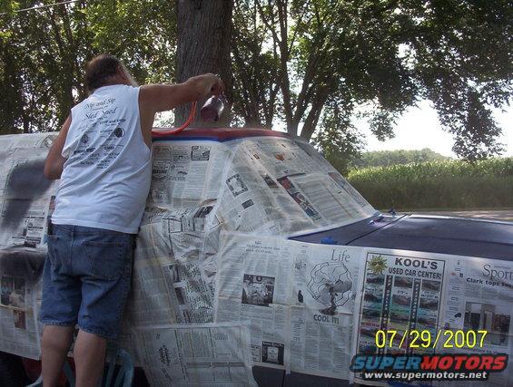 Man in denim shorts and white tank top sprays paint on a dark blue vehicle covered in newspaper