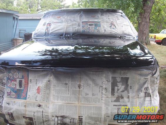 Front of a car covered in newspaper and plastic for painting. A yellow car is visible in the background