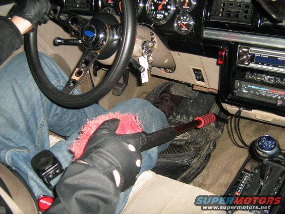 Person in jeans and black gloves cleaning car interior with a pink duster. Aftermarket steering wheel and radio visible