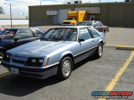 Light blue 1986 Ford Mustang coupe parked in a lot, with a black car and a brown sedan nearby