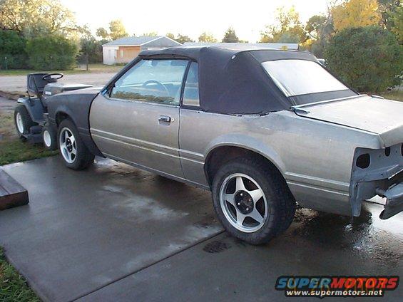 Silver 1984 Ford Mustang convertible side view with black top and a lawn tractor attached
