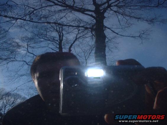 Person holding a digital camera with flash on, reflected in a dark surface