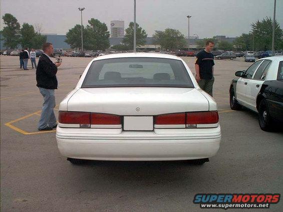 Rear view of a white sedan parked in a lot. A police car is visible to the right