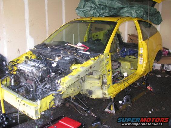 Yellow car with front end removed, engine exposed, doors open, and interior stripped. Car is on jack stands in a garage