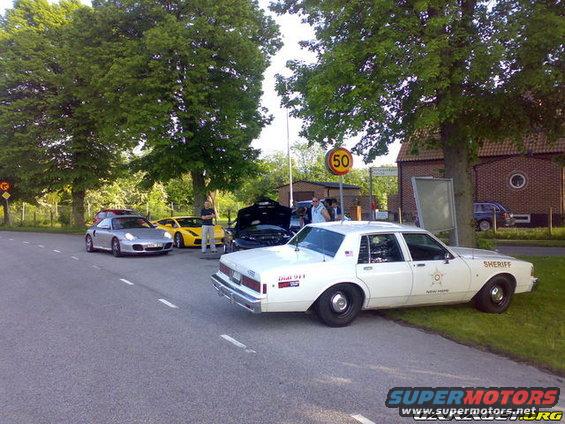 White sheriff car parked on grass, side view. Other sports cars visible