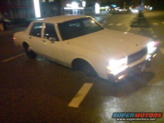 White sedan parked on wet street at night, headlights on