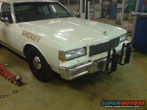 White sheriff's car with gold 'SHERIFF' text, push bumper, and chrome grille, viewed from a 3/4 front angle