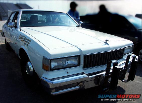 White sheriff car, likely 1980s model, with push bumper and 'SHERIFF' text on fender. Another dark vehicle is partially visible