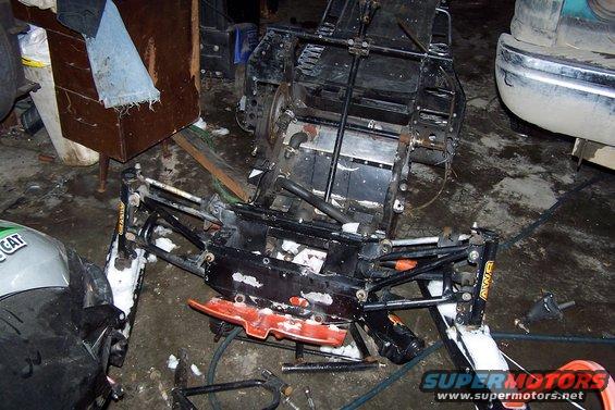 Snowmobile chassis and front suspension assembly, partially disassembled. A green and silver hood section with 'CAT' visible is on the left