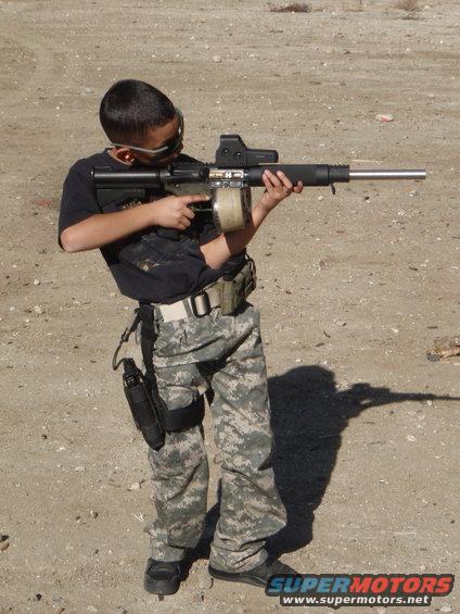 Kid in camo pants and tactical vest holding a rifle with a drum magazine and red dot sight