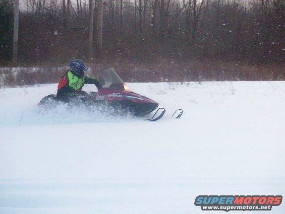 Red snowmobile with a rider in a yellow vest driving through snow, kicking up spray