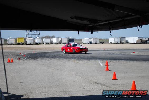 Red Ford Mustang with white racing stripes on a track, with orange cones and trailers in the background