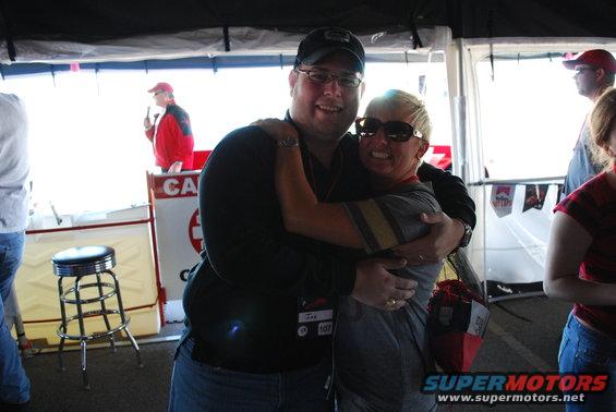 Two people embracing in front of a tent with a stool and a person in a red jacket in the background
