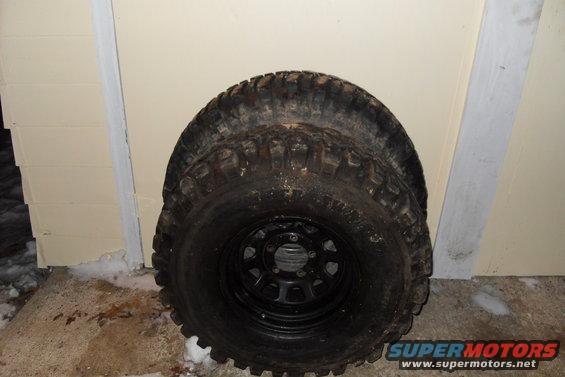 Two large off-road tires stacked on top of each other, leaning against a wall. One tire is on a black rim