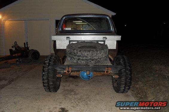 Rear view of a modified pickup truck with large tires, a spare tire mounted on the back, and a diamond plate toolbox
