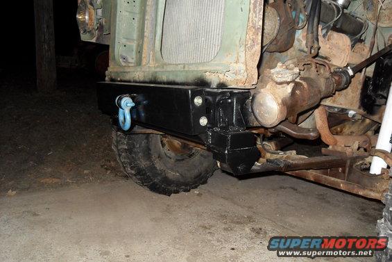 Front of a vehicle showing a black aftermarket bumper with a blue tow hook, part of the suspension, and the radiator