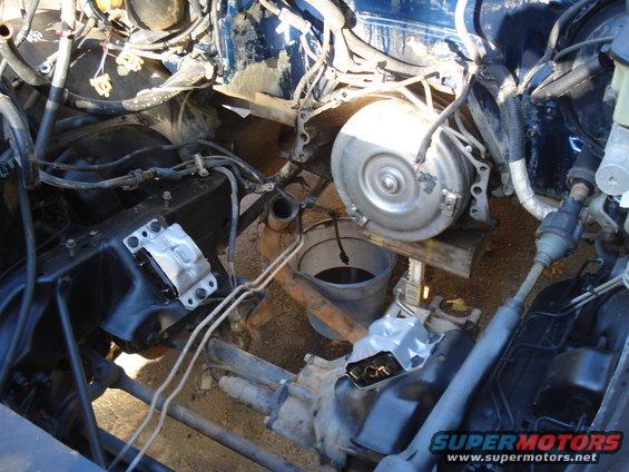 Vehicle engine bay with transmission removed, showing engine mounts and drivetrain components. A bucket of dark fluid sits nearby